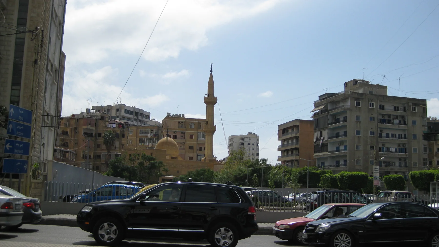 Cars pass in front of apartment buildings and a slender minaret beneath a partly cloudy sky.