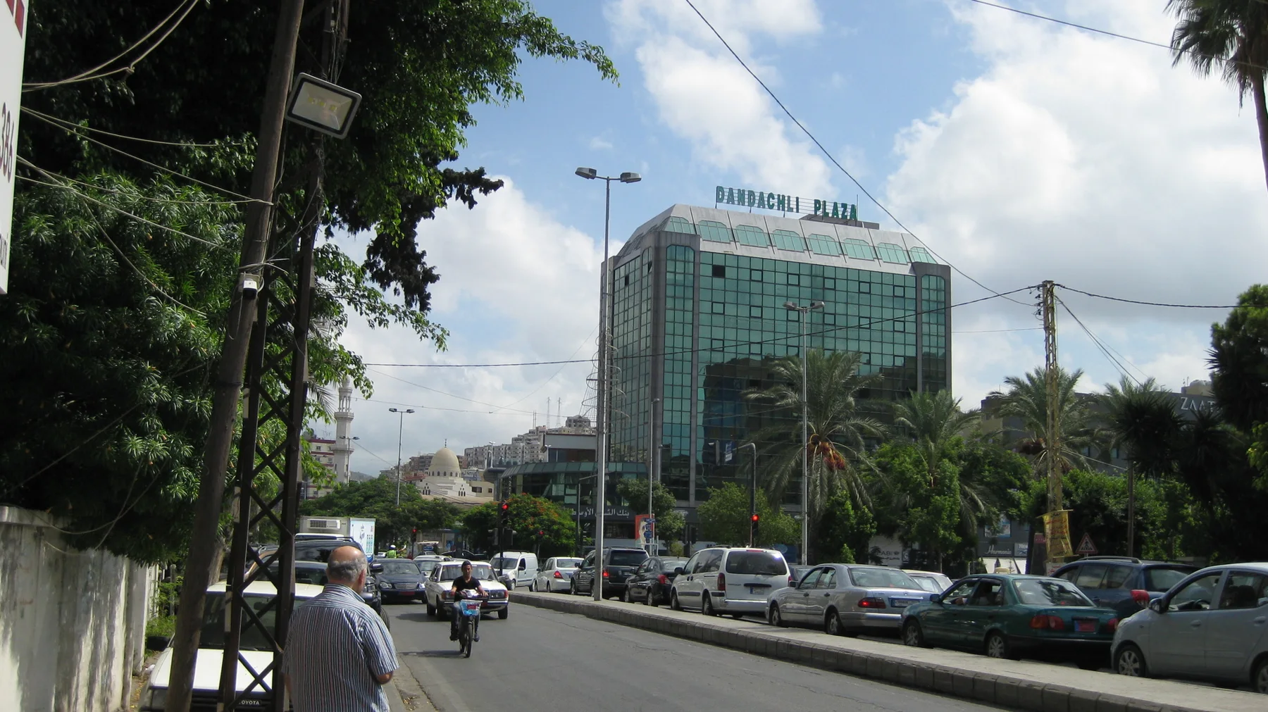 Traffic fills a broad road beside trees and a large glass-fronted building, with overhead cables crossing the view.