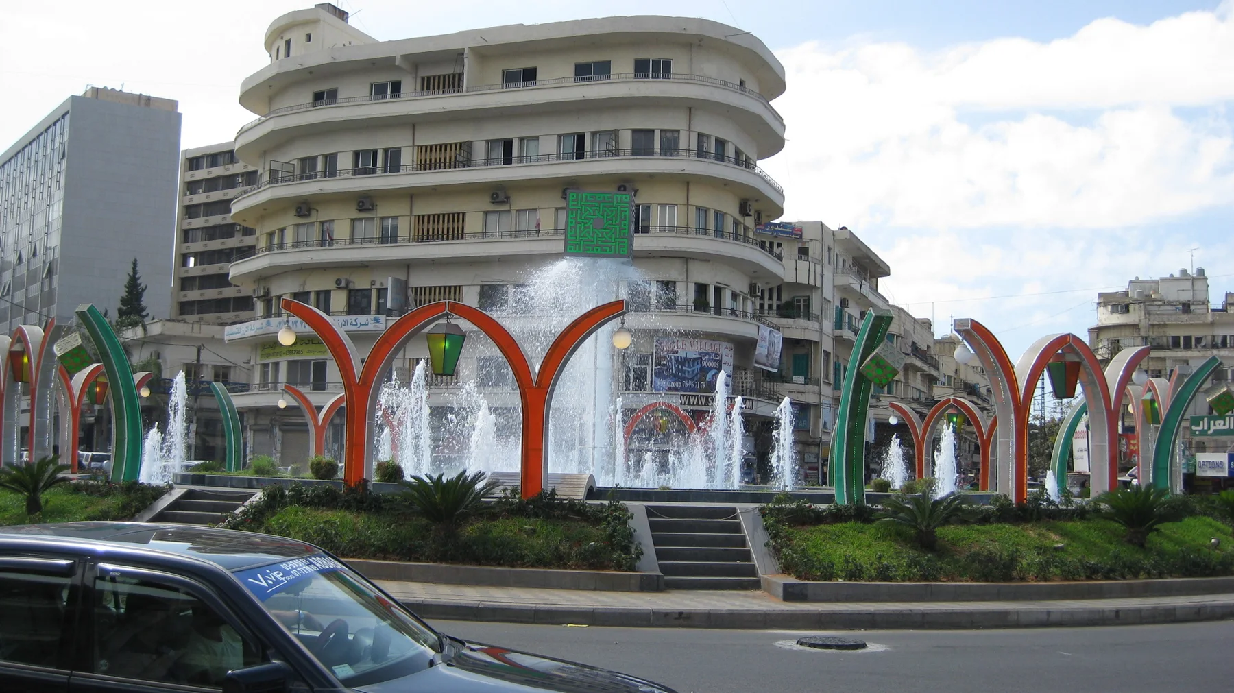 A fountain with colourful curved arches stands before a rounded white building, with passing cars in the foreground.