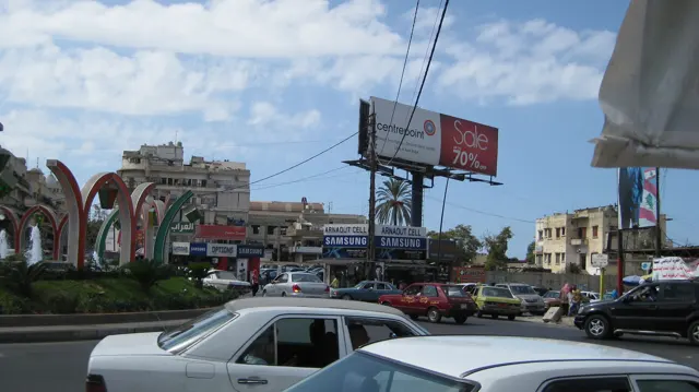 Cars crowd a junction near colourful fountain arches, roadside signs and overhead advertising.
