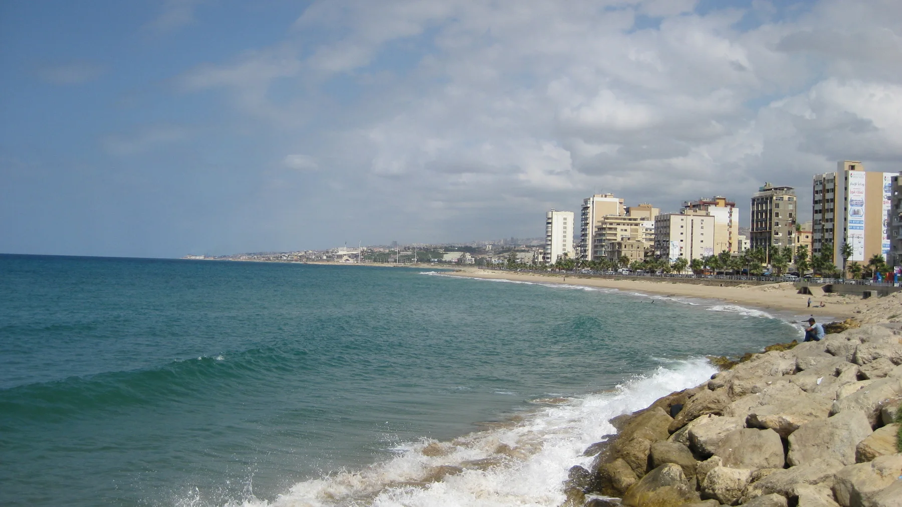 Blue-green water meets a curving shoreline lined with buildings, with large rocks in the foreground and clouds above.