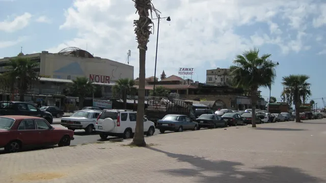 Parked cars line a broad roadside area beside low shops, palm trees and signs beneath a blue sky.