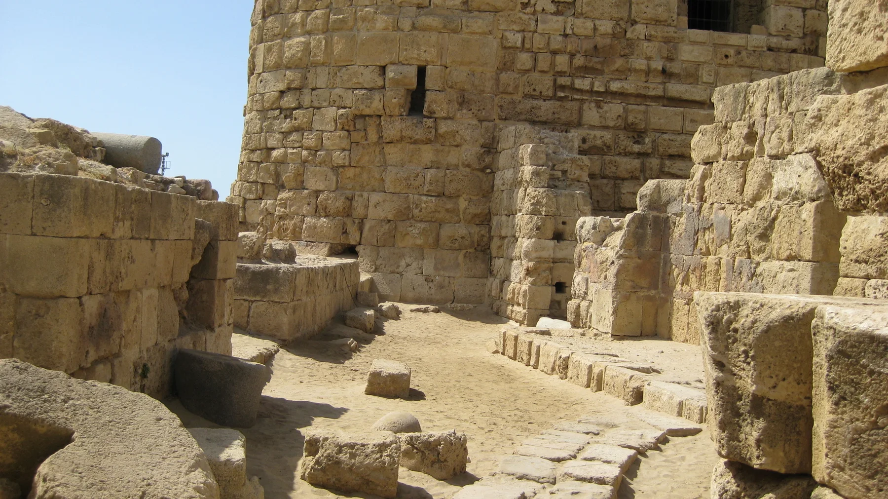 Sunlit stone blocks and low walls surround an open space beside a rounded tower built from pale masonry.