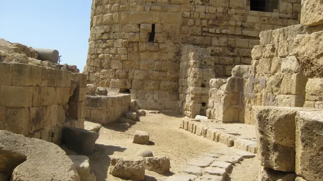 Sunlit stone blocks and low walls surround an open space beside a rounded tower built from pale masonry.