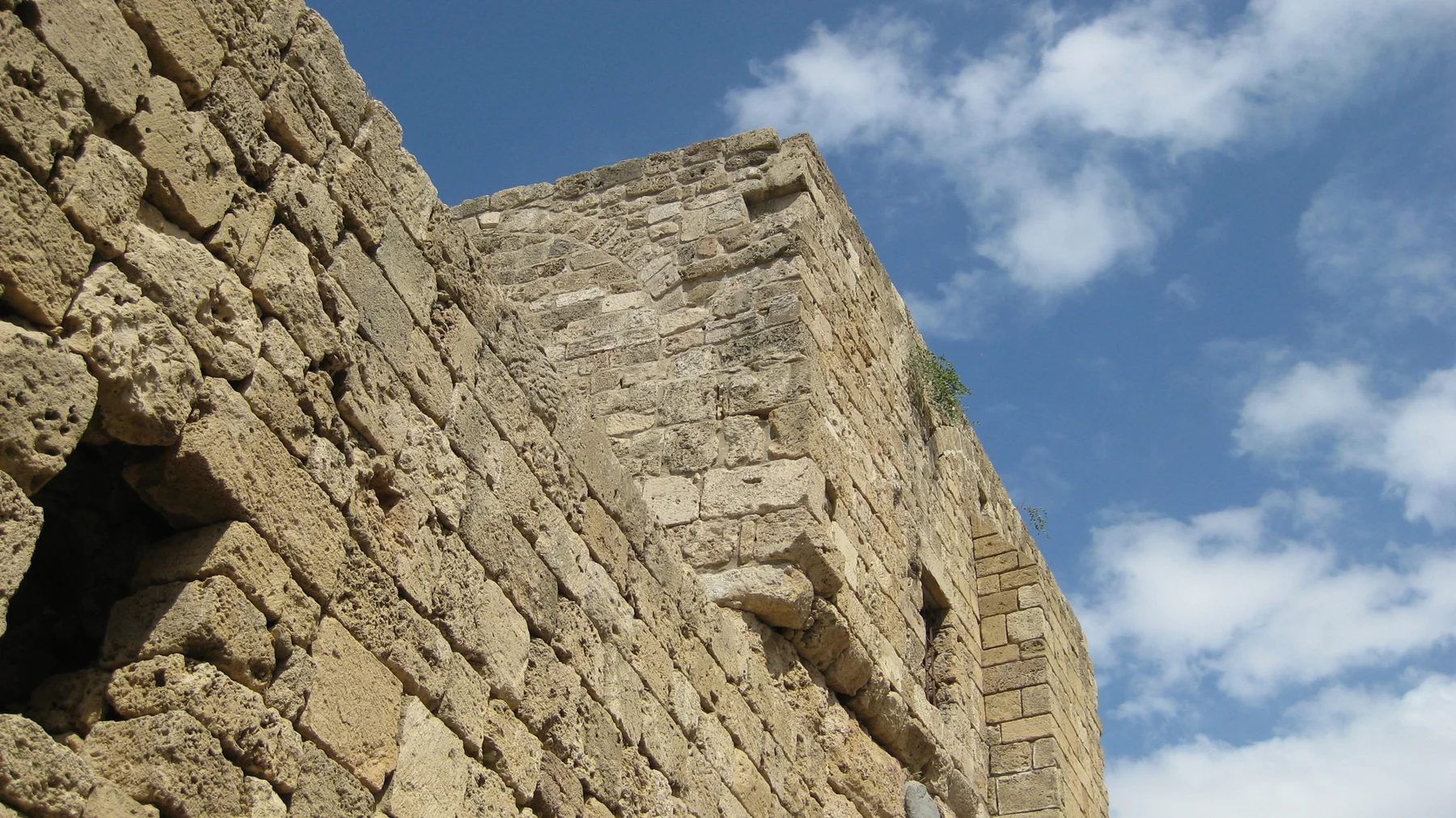 An upward view follows rough pale stonework and projecting blocks towards a blue sky with scattered clouds.