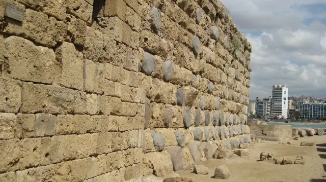 A long rough stone wall fills one side of the frame, with an open paved area and distant buildings beyond.