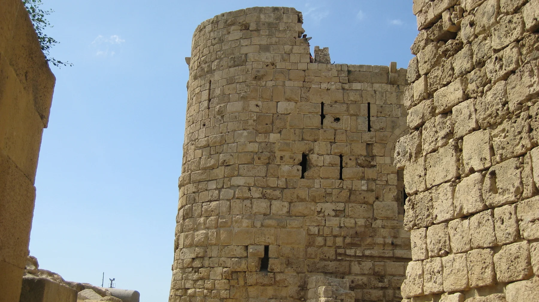 A rounded masonry tower with narrow openings rises between weathered walls against a clear blue sky.