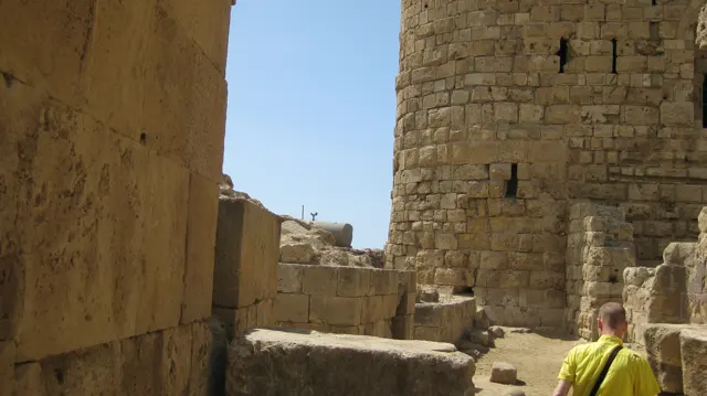 A visitor in a bright top walks through a sunlit space between massive stone walls and a rounded tower.