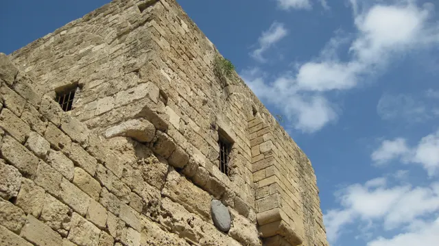 Small openings and projecting stone blocks punctuate a tall rough masonry wall beneath scattered clouds.