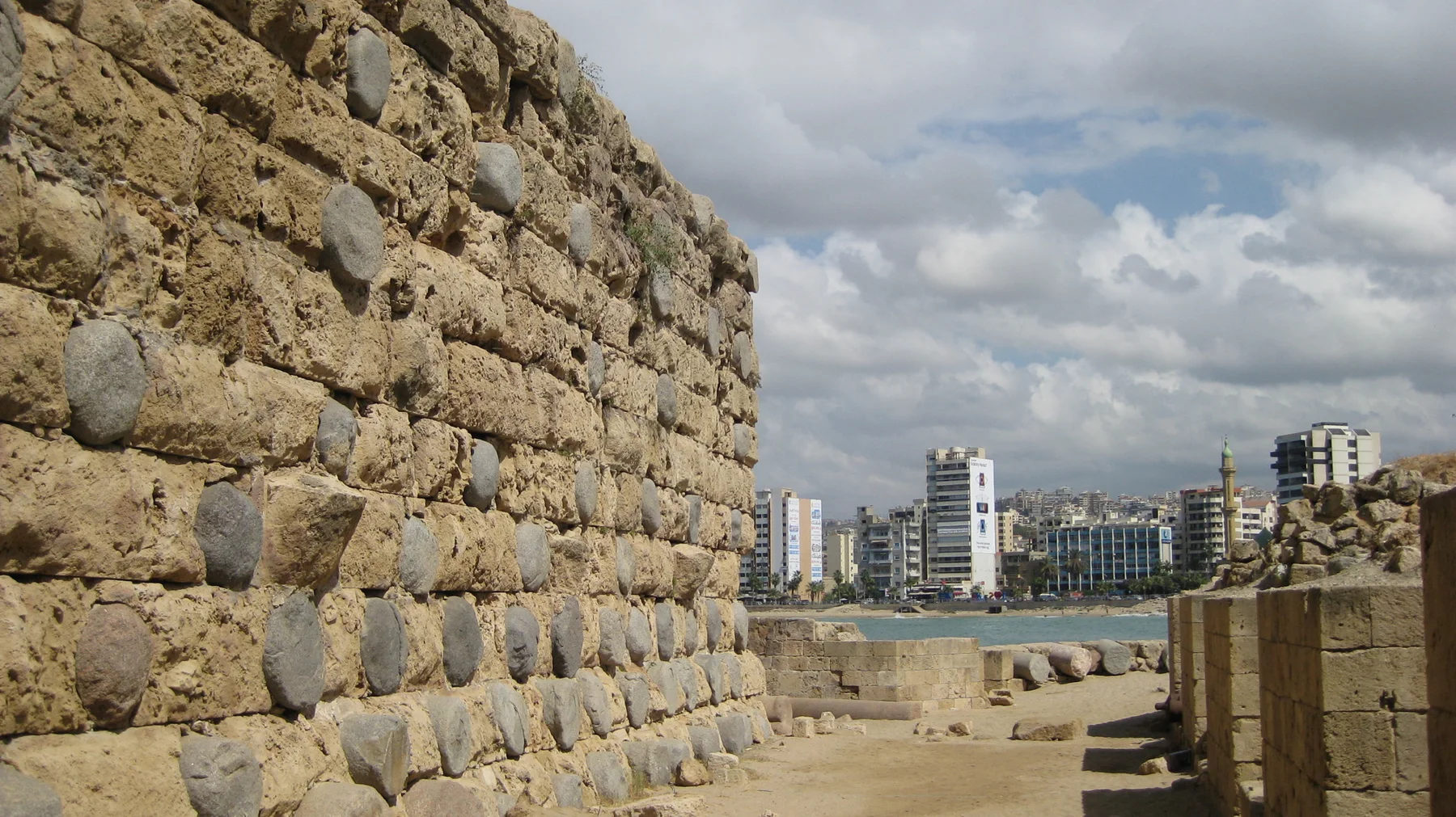 A weathered stone wall leads towards a view of water and distant waterfront buildings beneath clouds.