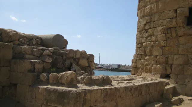 Rough low walls and scattered stone blocks border a rounded tower, with blue water visible through a gap.