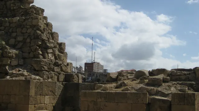 Uneven stone walls frame a view towards distant buildings and a tall mast beneath large clouds.