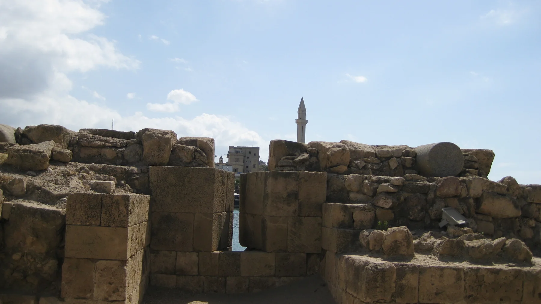 Low weathered walls and large stone blocks partly obscure a distant minaret beneath a pale blue sky.