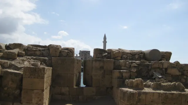 Low weathered walls and large stone blocks partly obscure a distant minaret beneath a pale blue sky.