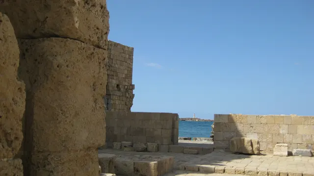 Thick stone walls and a low parapet frame a narrow view of blue water under a clear sky.
