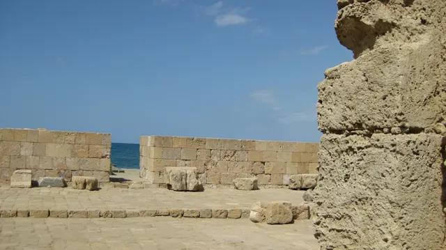 A gap in a low stone wall opens towards blue water, with large blocks scattered across a sunlit paved area.