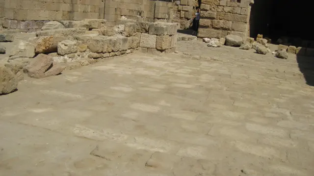 A broad worn stone surface fills the foreground of a courtyard bordered by masonry, loose blocks and a dark doorway.