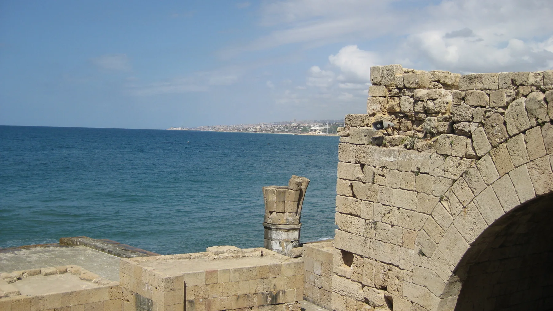 Part of a stone arch and parapet stands above blue water, with a slim stone feature near the edge.