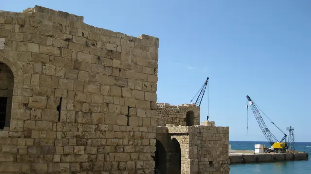 Weathered masonry buildings stand beside the sea, with tall harbour cranes visible beyond.