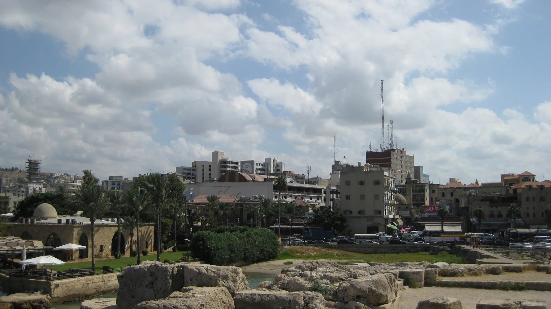 Stonework in the foreground overlooks a waterfront road, trees and closely packed buildings beneath a cloud-filled sky.