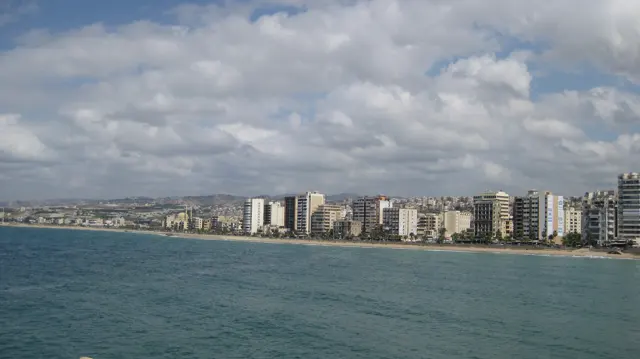 A long line of waterfront buildings follows the shore across blue-green water beneath banks of white clouds.