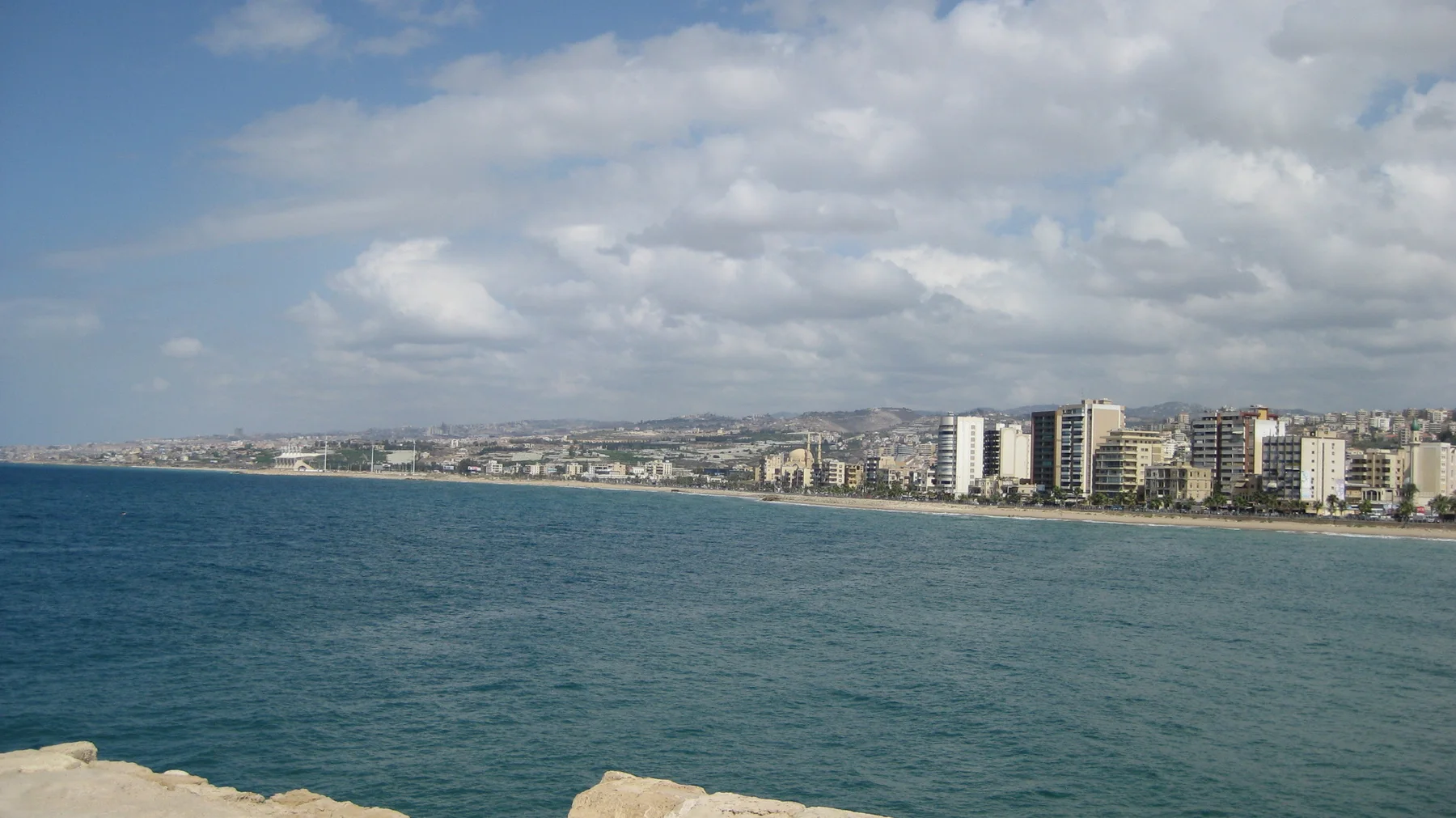 A waterfront skyline curves around the bay beyond deep blue water, with pale stone at the lower edge.
