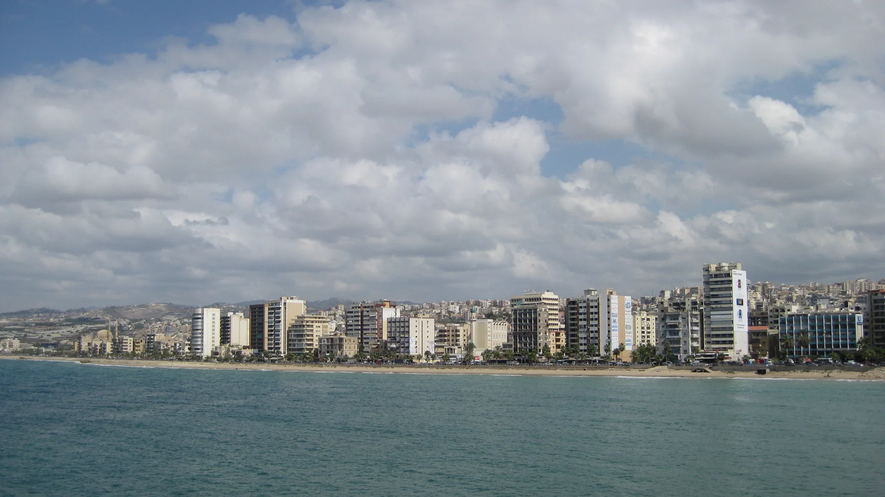 Closely spaced buildings face the water beneath scattered clouds in a broad coastal view.