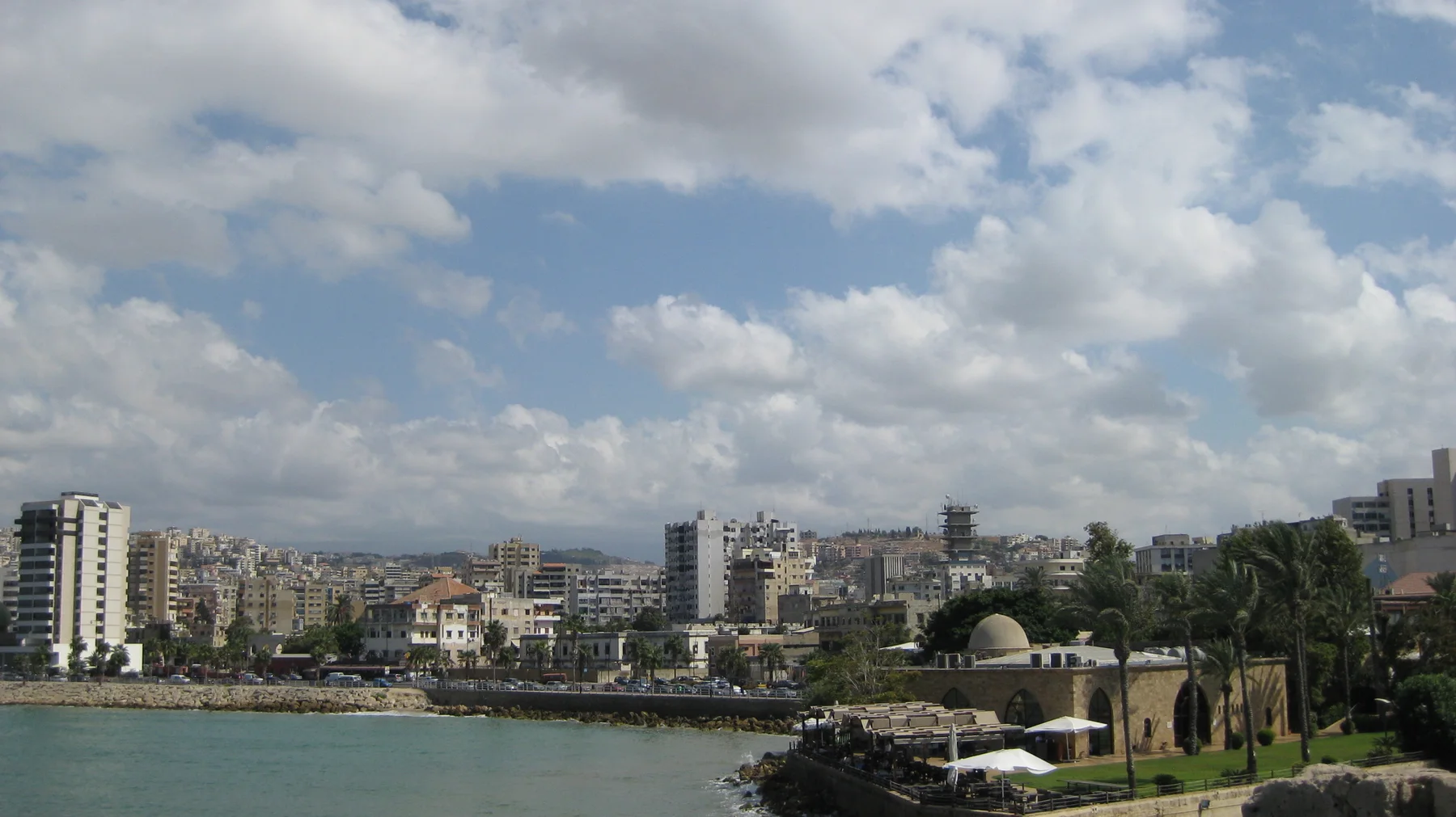 A small domed waterfront building and trees stand before a dense townscape across the bay.