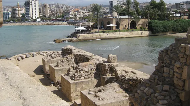 Low ruined walls and passages lie below an elevated viewpoint, with water and the town beyond.