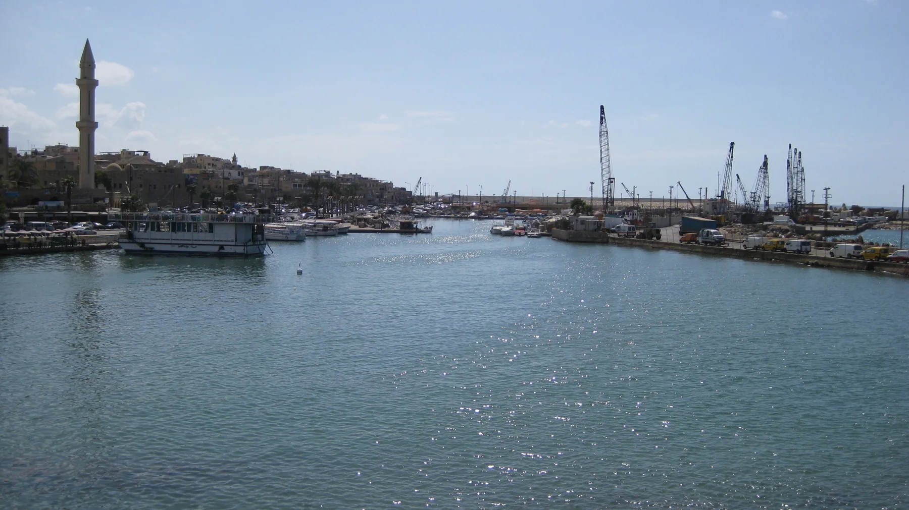 Boats and harbour cranes line a sheltered stretch of water, with a slender minaret at the left.