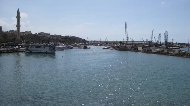 Boats and harbour cranes line a sheltered stretch of water, with a slender minaret at the left.