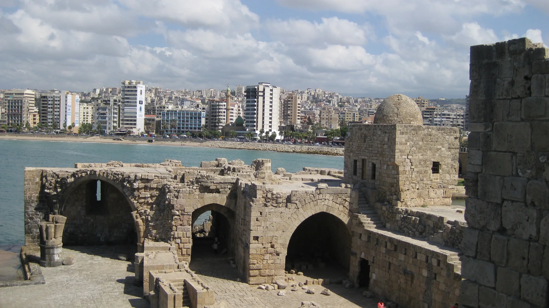 Several dark arched openings and a small dome stand among pale stone walls, with waterfront buildings beyond.