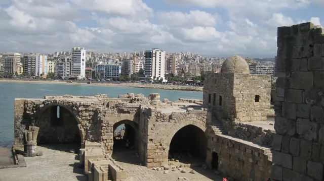 Several dark arched openings and a small dome stand among pale stone walls, with waterfront buildings beyond.