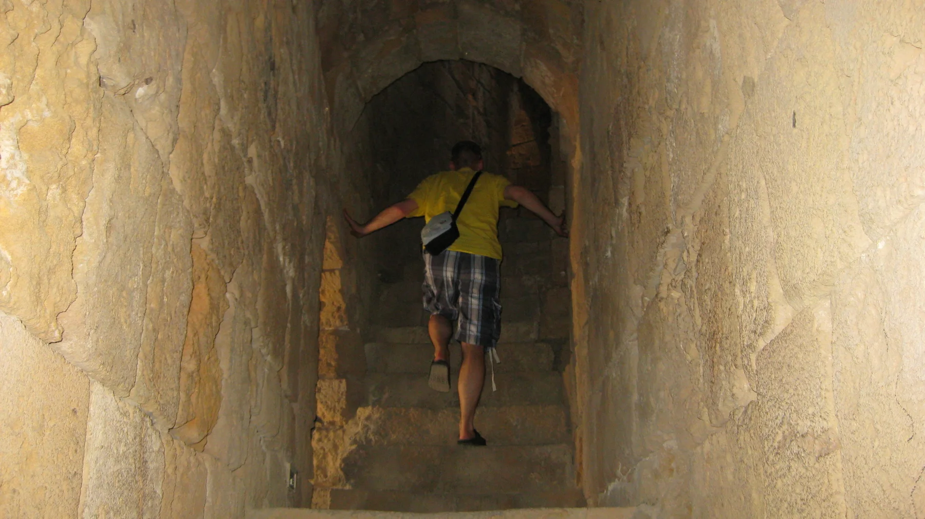 A visitor in a yellow top descends a narrow stairway between thick stone walls beneath an arch.