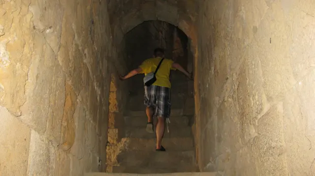 A visitor in a yellow top descends a narrow stairway between thick stone walls beneath an arch.