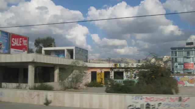 A low unfinished-looking building and roadside signs pass beneath large clouds, with taller structures nearby.