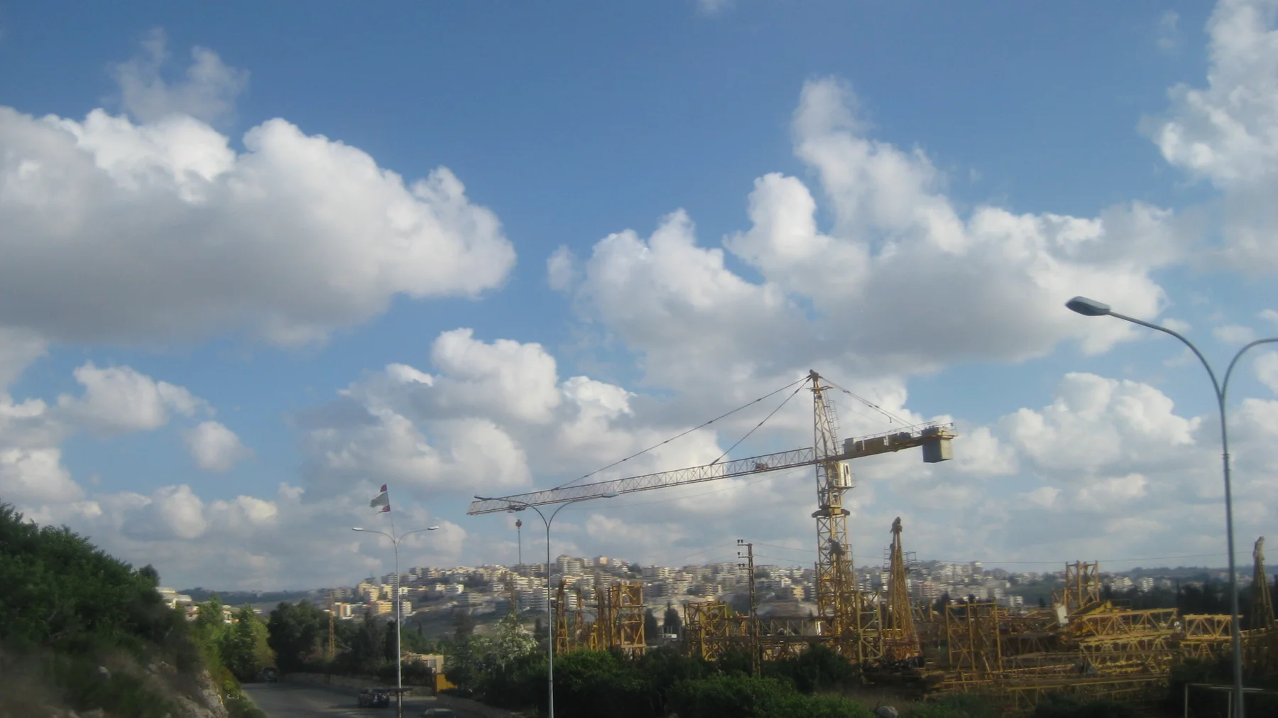 A construction crane stretches across the skyline above low structures beneath large white clouds.