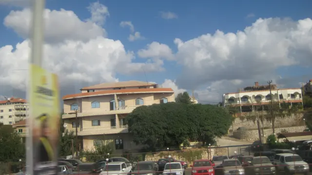 Low buildings with balconies and tiled roofs stand behind cars beside a roadside sign.