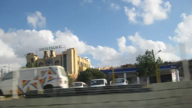 A large yellow-toned building and smaller commercial structures stand beyond passing traffic and a roadside barrier.