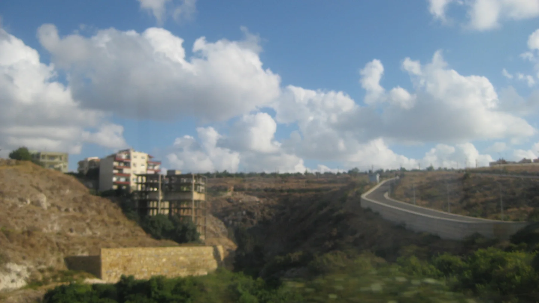 A paved road curves over a dry hillside beside scattered apartment blocks and exposed earth.