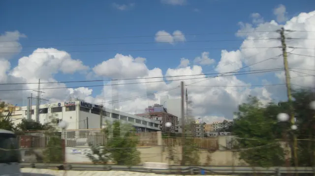 Utility poles and numerous wires cross a view of unfinished buildings and vegetation.