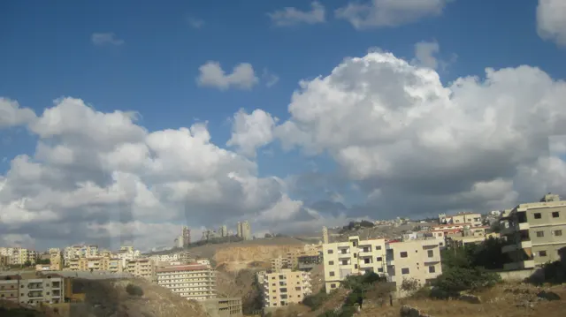 Pale apartment blocks line a dry hillside beneath a deep blue sky and large white clouds.