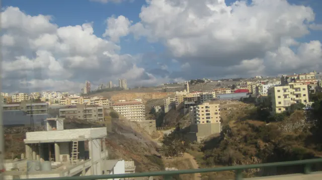 Apartment blocks and exposed rock cuts stretch across a sloping landscape under broken clouds.
