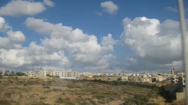 Low scrub and dry ground extend towards a line of pale buildings on the horizon.