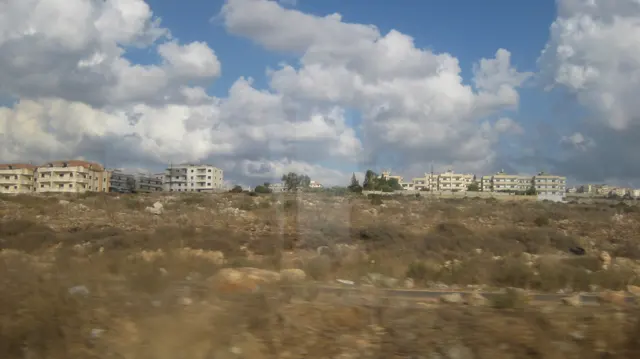 Scattered buildings stand beyond dry scrub and rough ground beneath bright white clouds.