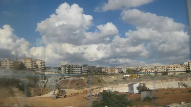 Excavated earth and pale rock lie beneath rows of hillside buildings in a view from the road.