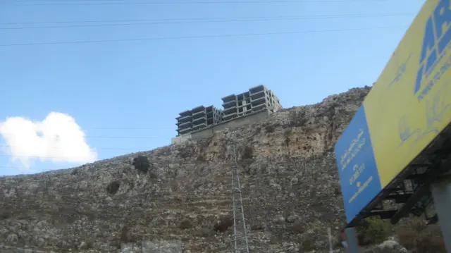 A high rough stone retaining wall rises beside a partly visible roadside sign under a blue sky.