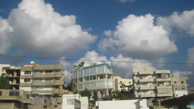 Several buildings with balconies and broad windows stand beneath large clouds and overhead wires.