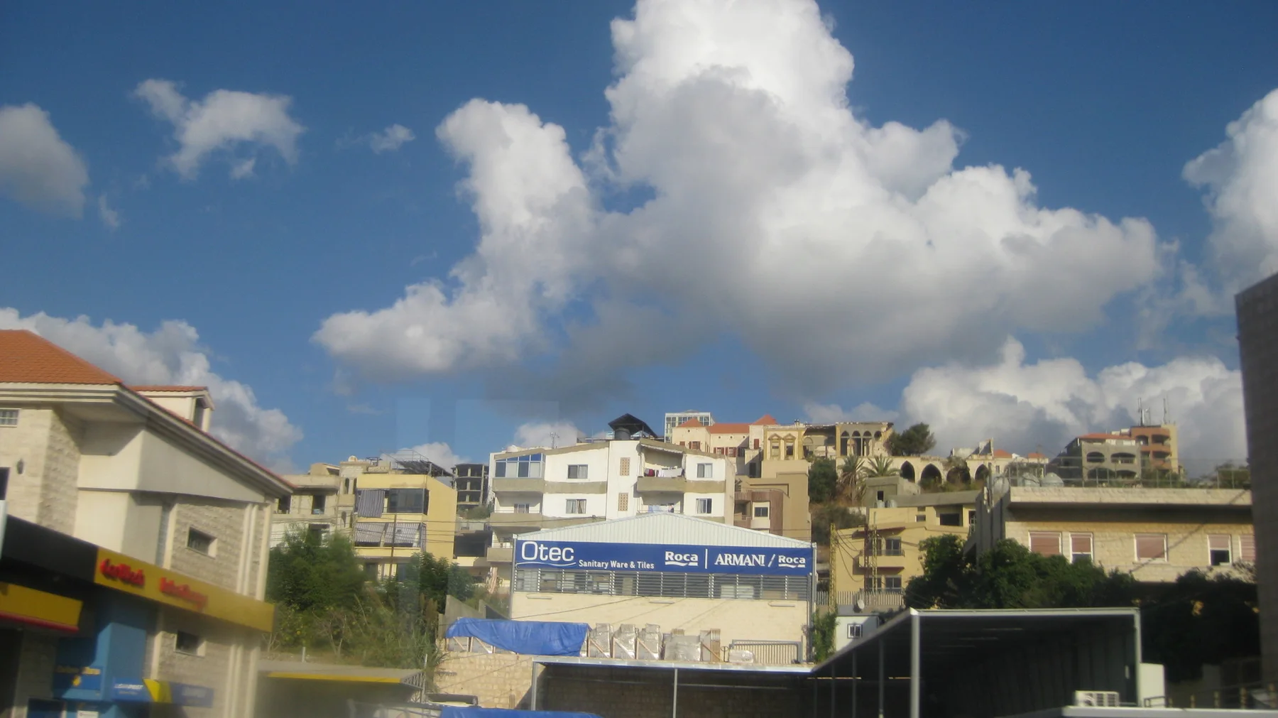 Roadside signs and low structures sit beneath densely arranged apartment blocks on a slope.