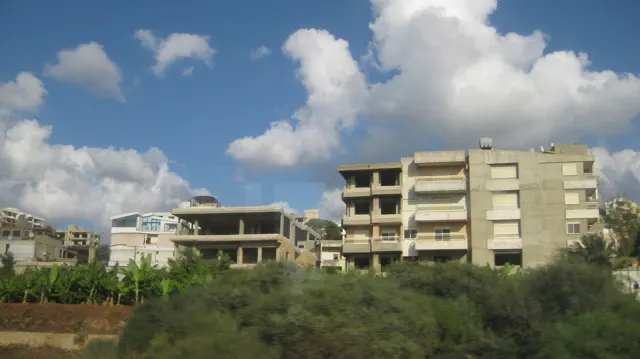 Apartment blocks and a partly open concrete structure rise beyond a patch of greenery.